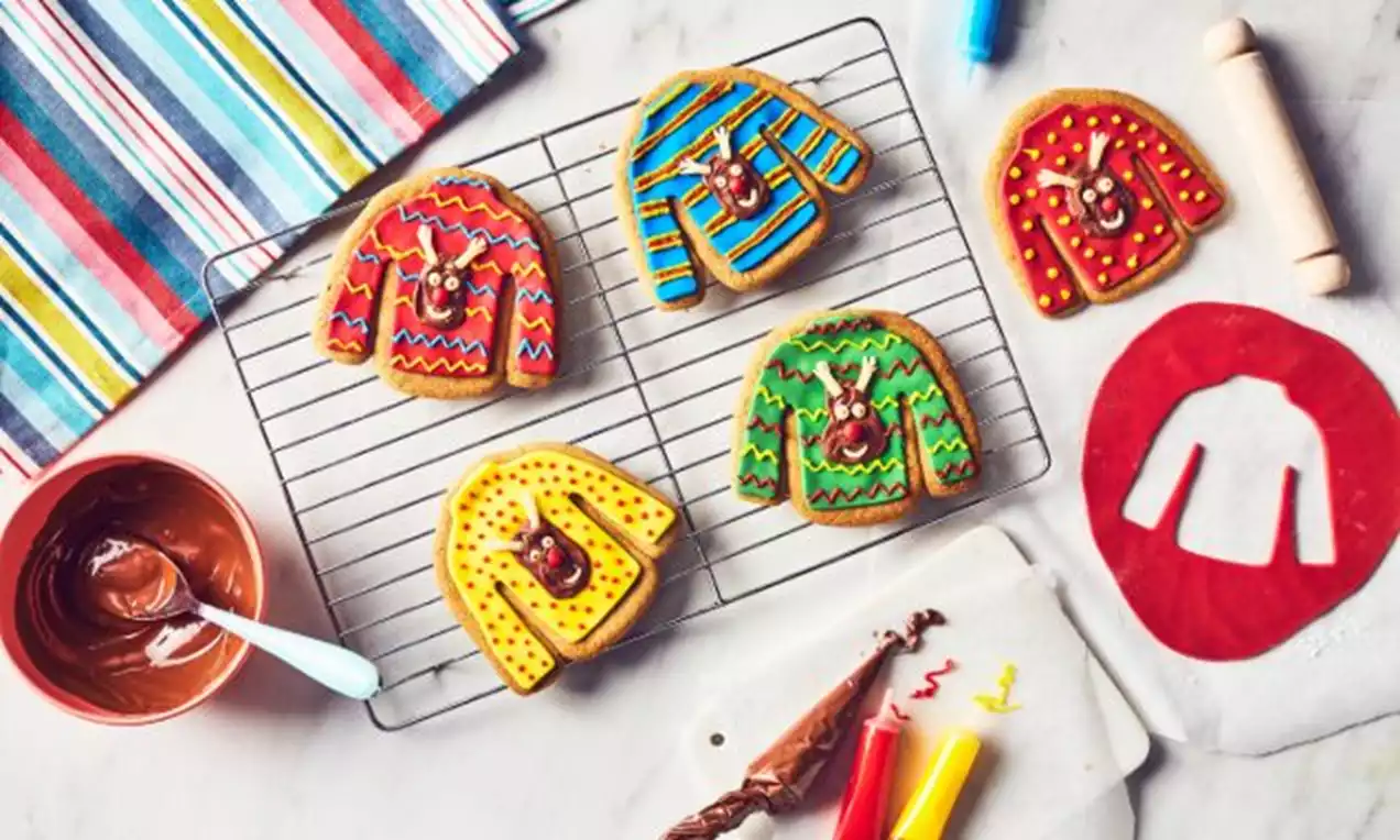 Christmas jumper-shaped biscuits iced in bright red, yellow, blue, and green patterns with reindeer faces on a cooling rack and marble surface